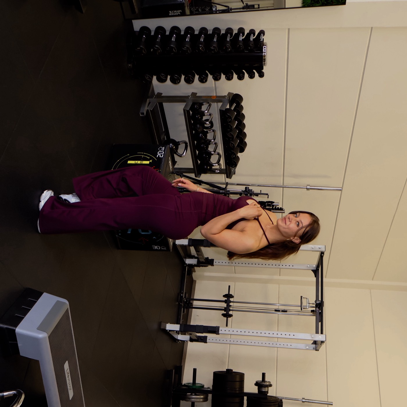 Fitnesscoach in bordeauxrode jumpsuit kijkt recht in de camera in een lichte gym
