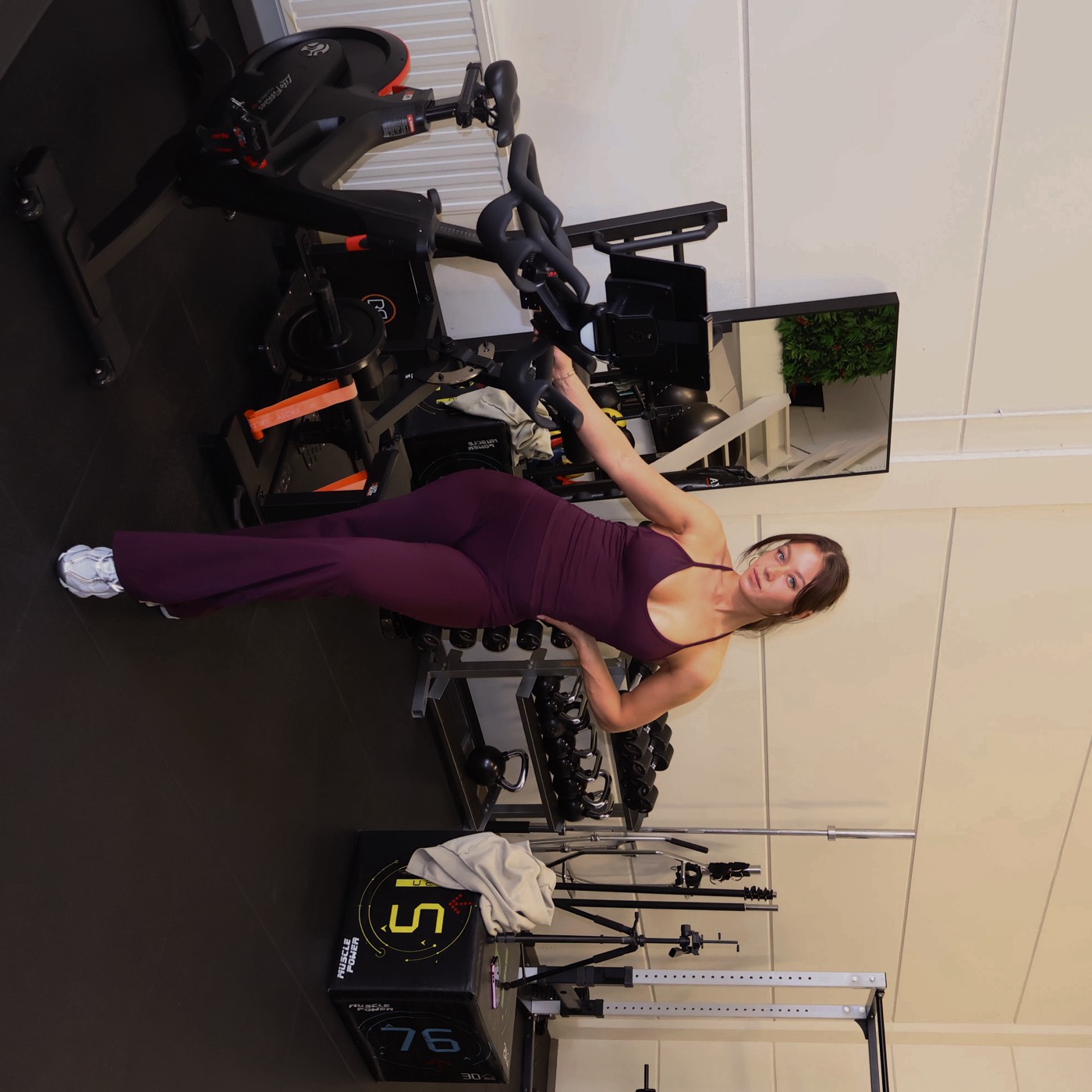 Vrouw in bordeauxrode jumpsuit leunt op een spinningfiets in een lichte gym