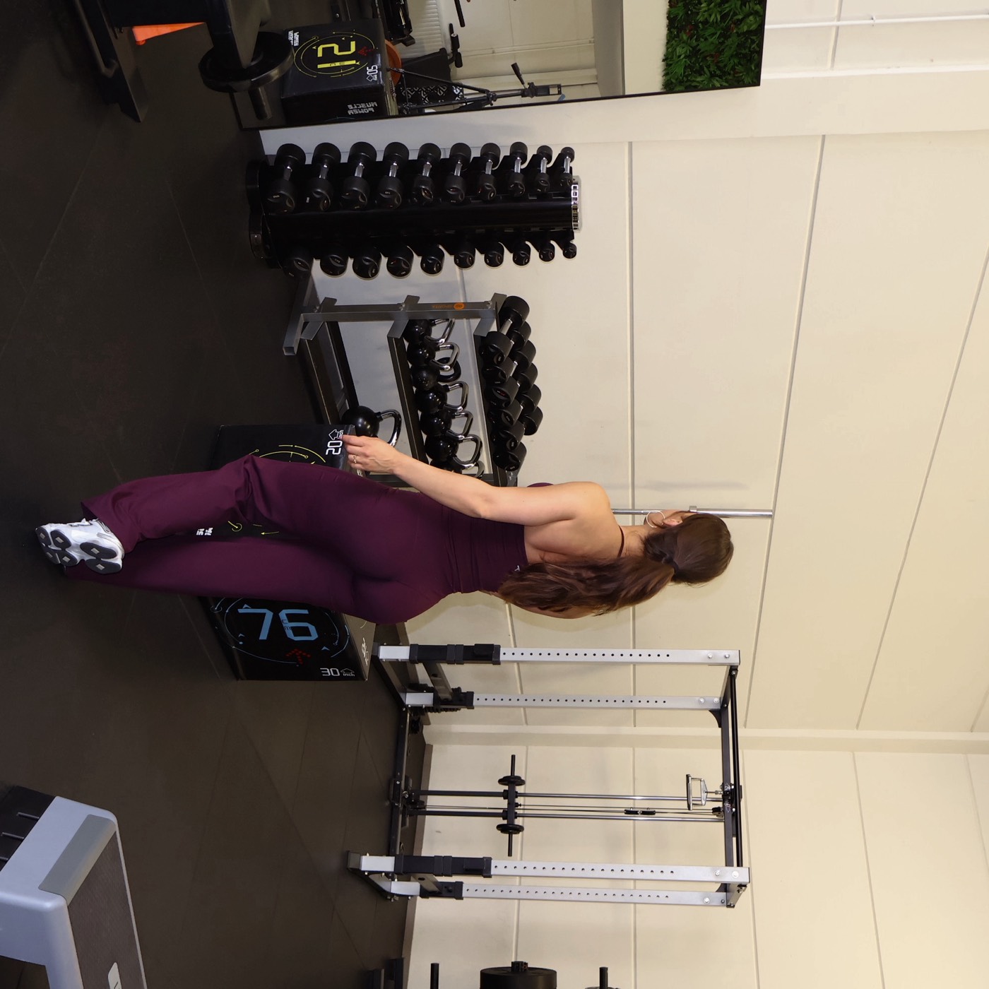 Vrouw in bordeauxrode jumpsuit bij een rek met dumbbells in een lichte gym