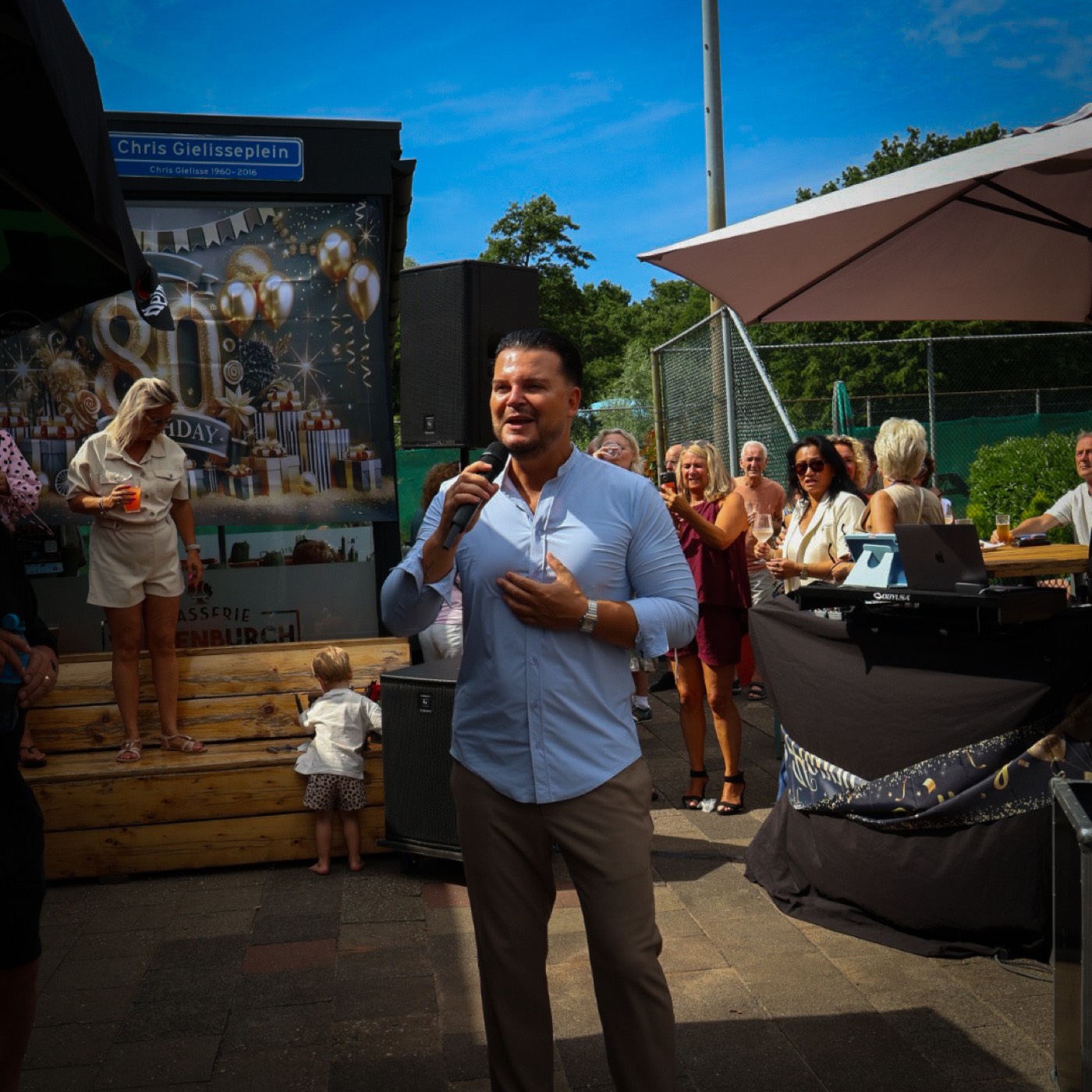 Mike Peterson zingt met microfoon in de hand op een zonnig terrasfeest met publiek